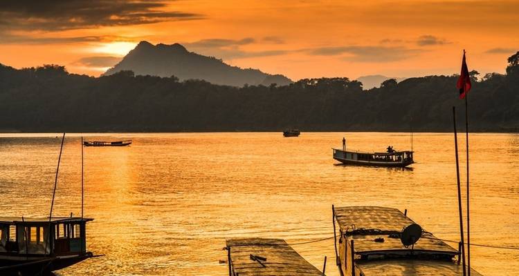Scenic river view at sunset with silhouettes of boats and mountains in the background.