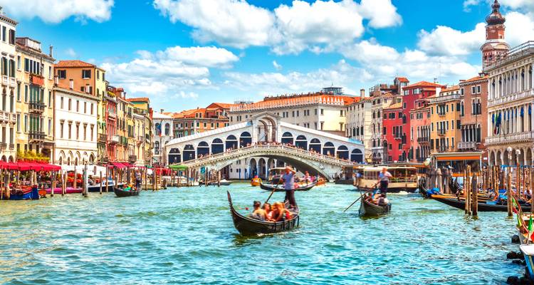Busy Grand Canal in Venice with gondolas and colorful historic buildings leading to the Rialto Bridge under a bright sky.