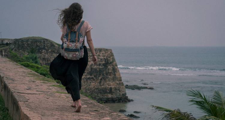 Femme pieds nus avec sac à dos marchant le long des murs historiques d'un fort maritime au crépuscule.