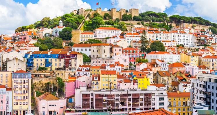 Farbenfrohe Häuser an Hängen und Burgmauern im Alfama-Viertel von Lissabon, gekrönt durch das Schloss São Jorge unter einem strahlend blauen Himmel.