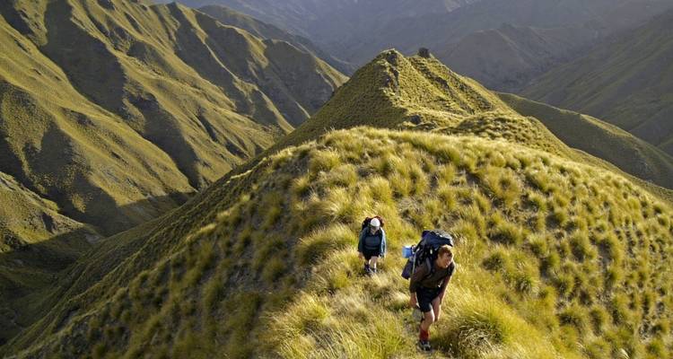 Two hikers climbing grassy hills in a mountainous landscape.