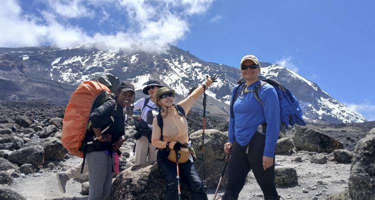 Quatre randonneurs posant sur une plaine rocheuse avec le sommet enneigé du Mont Kilimandjaro sous un ciel bleu dégagé.