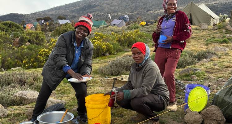 Trois femmes dans un camping de montagne lavant la vaisselle à côté de seaux colorés et de tentes sur un terrain herbeux.