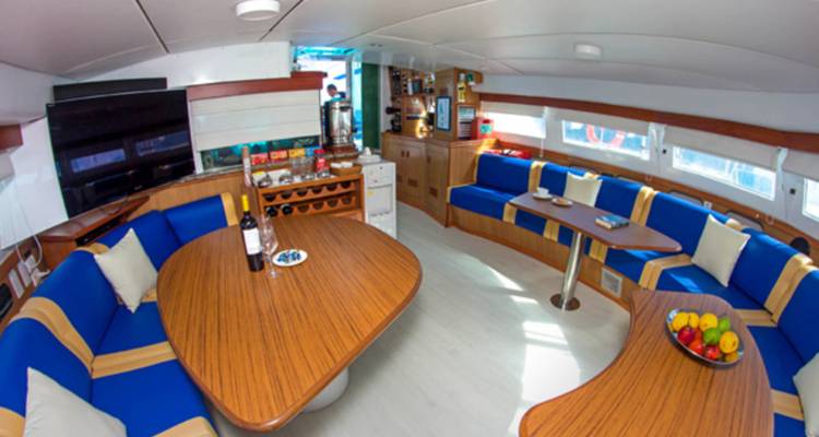 Acogedor salón de yate con mesas de madera con vino, un televisor de pantalla plana y asientos azules curvados mientras un miembro de la tripulación trabaja en el fondo.