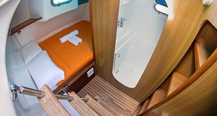 Camarote compacto de yate con cama cubierta en naranja, pequeña ventana y baño privado contiguo.
