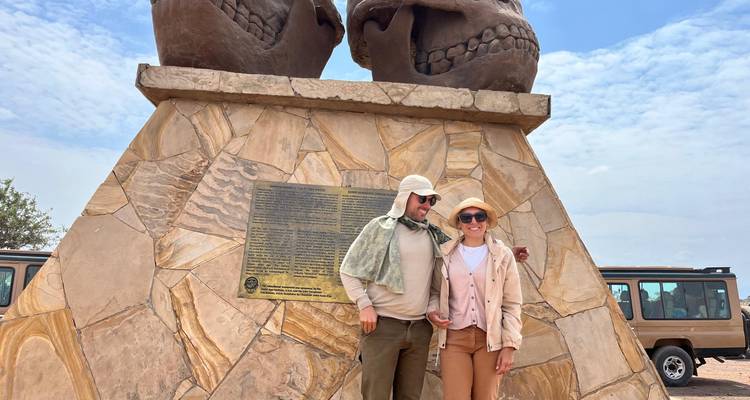 Couple poses in front of a large stone monument of two skulls near safari vehicles under a bright sky.