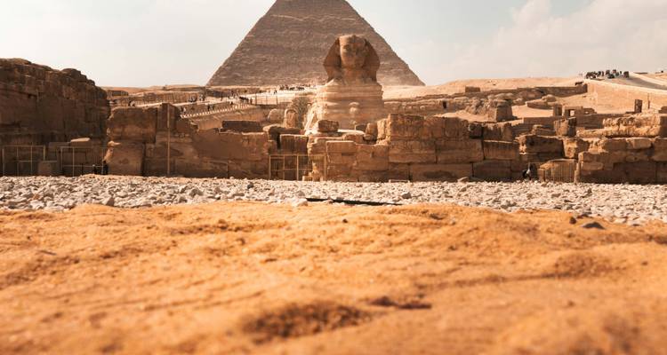 La Esfinge se encuentra frente a una pirámide enmarcada por arena dorada del desierto bajo un cielo pálido.