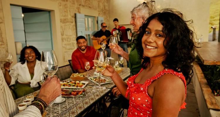 Des voyageurs souriants lèvent des verres de vin blanc autour d'une table à manger en plein air couverte de plats de tapas sur une terrasse en pierre.