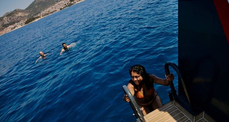 Femme en bikini montant une échelle de bateau sortant d'une mer bleu profond tandis que deux compagnons nagent à proximité avec des montagnes côtières en arrière-plan.