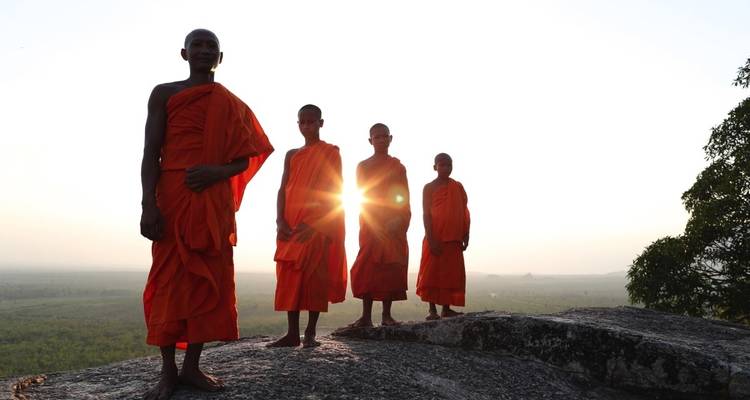 Vier Mönche in traditionellen orangefarbenen Roben stehen auf einem felsigen Vorsprung mit einer weiten Landschaft hinter ihnen.