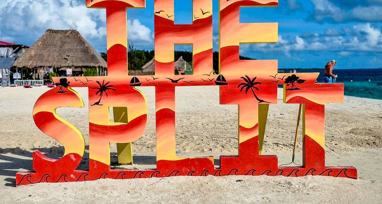 Un colorido letrero de playa que dice 'THE SPLIT' se encuentra en la arena blanca con agua turquesa y cielo azul.