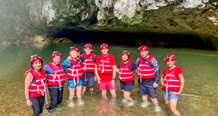 Un grupo con cascos se para hasta los tobillos en un río dentro de una caverna usando chalecos salvavidas rojos antes de hacer tubing en la cueva.