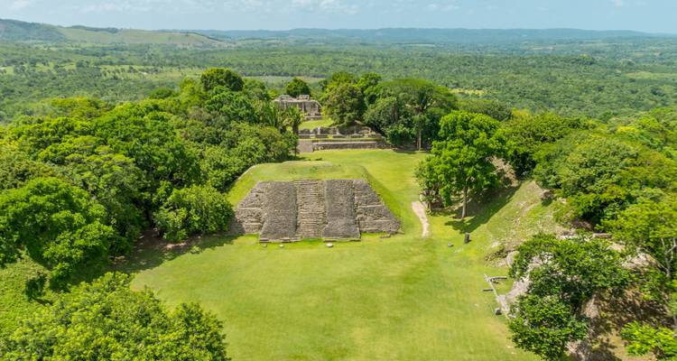 Aerial view of ancient Mayan plazas and pyramid ruins surrounded by dense green jungle hills.