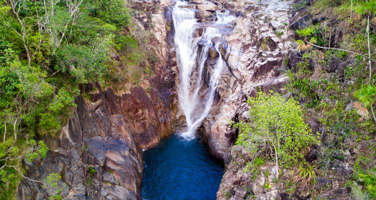 Aerial view of a tall waterfall plunging into a deep blue pool, surrounded by rugged rock cliffs and lush greenery.