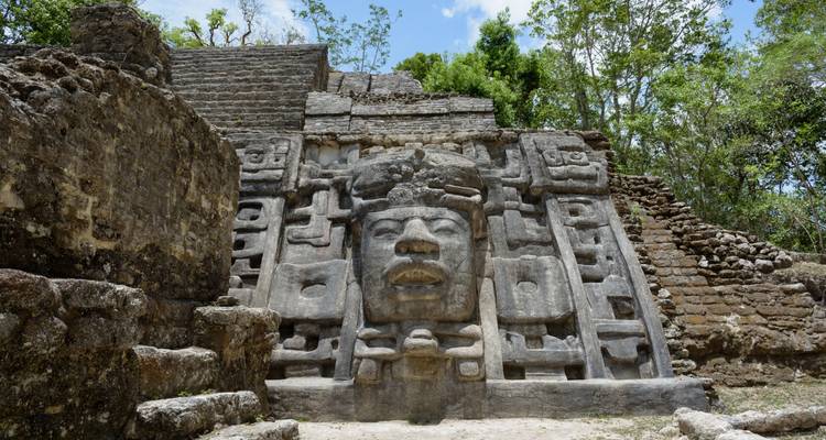 Close-up of an intricately carved Mayan stone mask façade at an ancient temple ruin set in jungle surroundings.