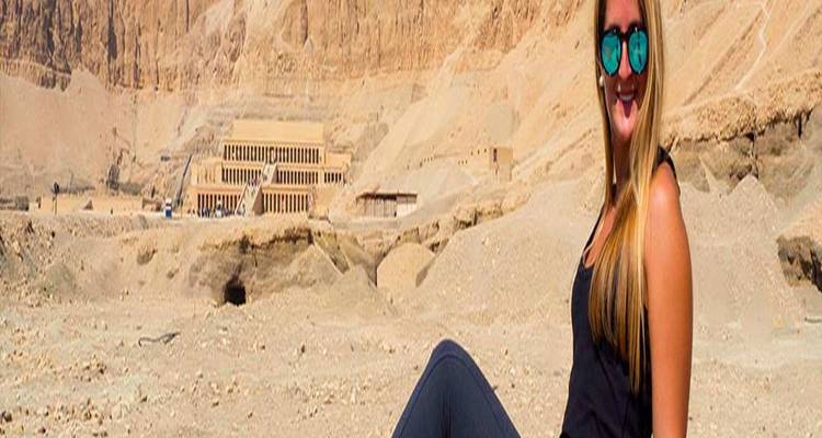 Mujer joven sentada contra un telón de fondo de desierto rocoso y estructuras antiguas.