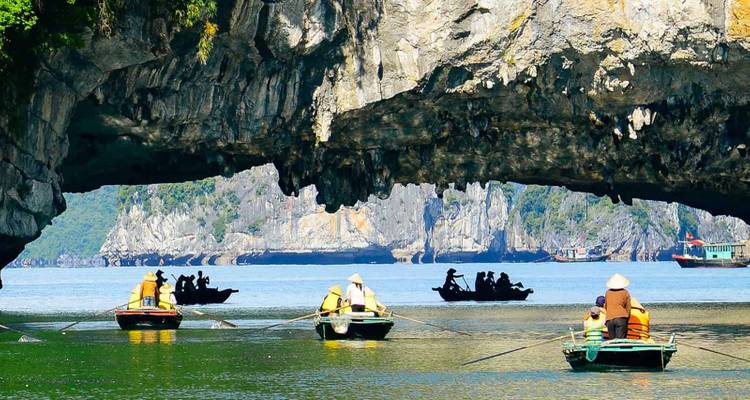 Groups of tourists rowing boat tours under a large limestone arch in a bay.