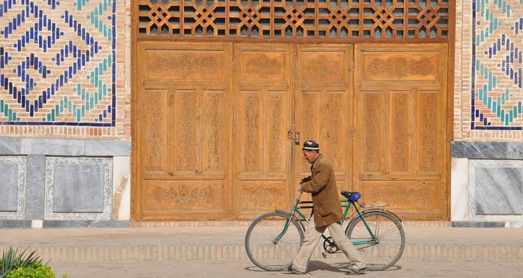 A man walking a bicycle past an ornate wooden door with mosaic details.