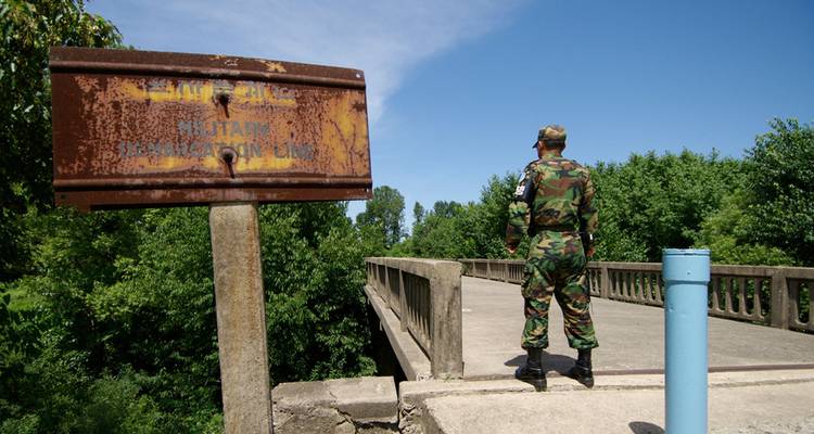 Uniformed soldier guarding a concrete bridge near a rusted military demarcation line sign on a sunny day.