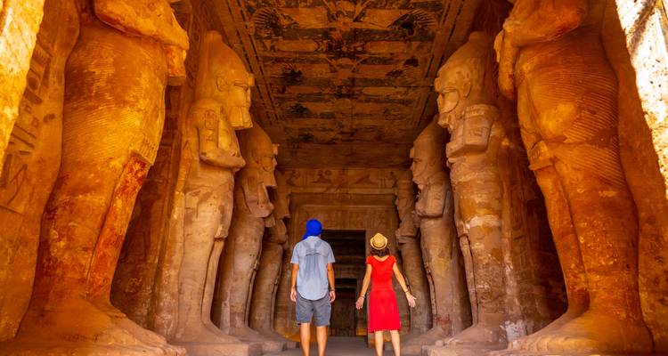 Two travelers stand between towering statues inside the grand rock-cut halls of Abu Simbel Temple bathed in warm light.