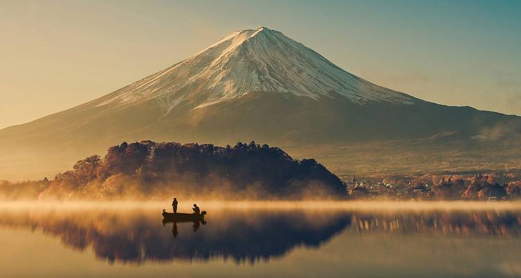 A snow-capped Mount Fuji rises majestically over a mist-covered lake where two people paddle a small boat at dawn.