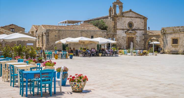 Las coloridas mesas de la terraza al aire libre iluminan una histórica plaza siciliana bañada por el sol, junto a una antigua iglesia de piedra.