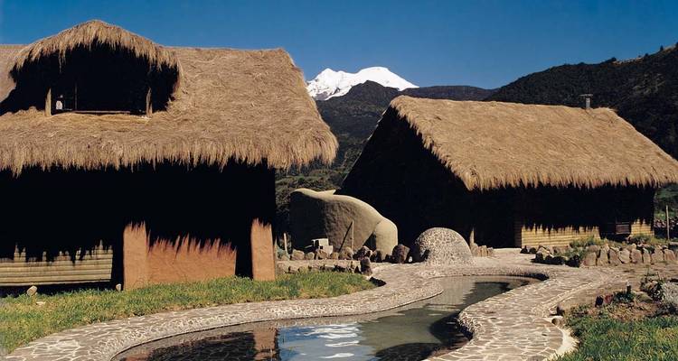 Thatched-roof lodges and a curving stone pool sit beneath a snow-capped volcano against a clear blue sky.