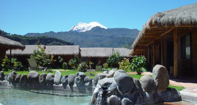 Rustic lodge buildings with thatched roofs overlook a tranquil stone pool with a snow-capped volcano in the distance.