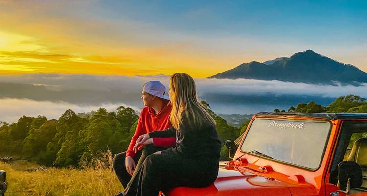 Dos personas se sientan en un Jeep rojo contemplando una vista de montaña al amanecer.