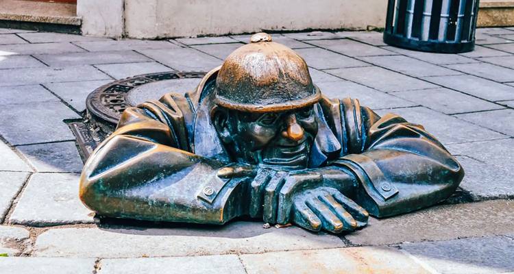 Bronzen standbeeld dat uit een putdeksel op straat omhoog komt.