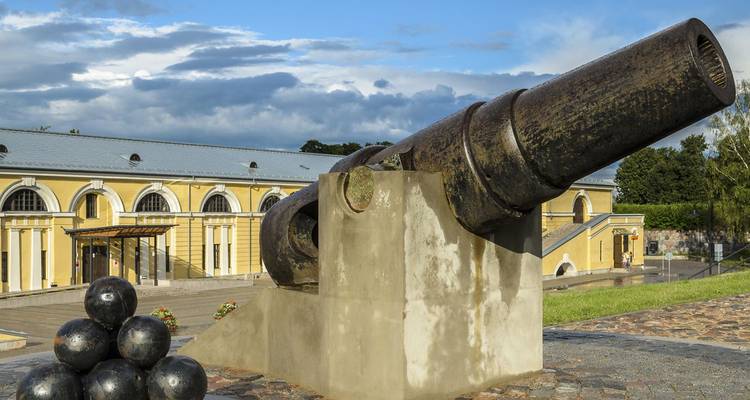 Historic black cannon and stacked cannonballs displayed outside a yellow arsenal building.
