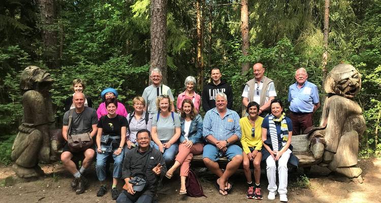 Tour group poses on wooden sculptures in a dense Baltic forest clearing.