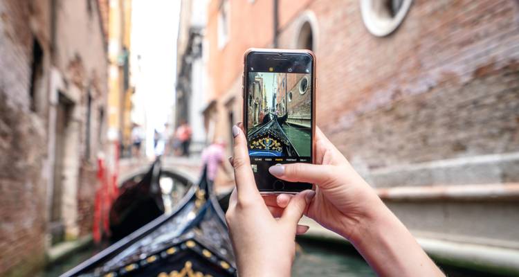 Gros plan des mains d'un voyageur tenant un téléphone et photographiant une promenade en gondole dans un canal étroit.