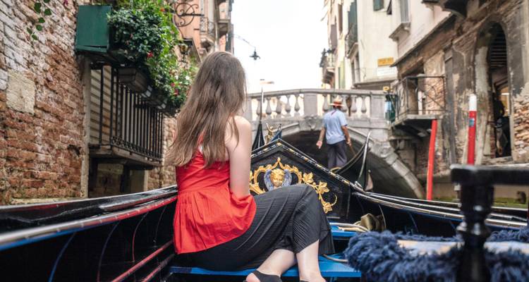 Femme vêtue d'un haut rouge assise à l'intérieur d'une gondole passant sous un petit pont en pierre.