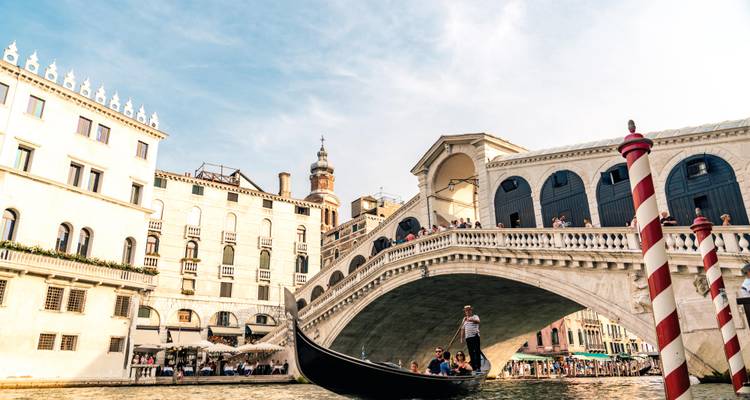 Escena clásica de una góndola con pasajeros pasando bajo el Puente de Rialto contra un cielo azul.