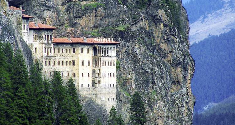 A historic monastery clings dramatically to a sheer forested cliff amid rugged mountain scenery.