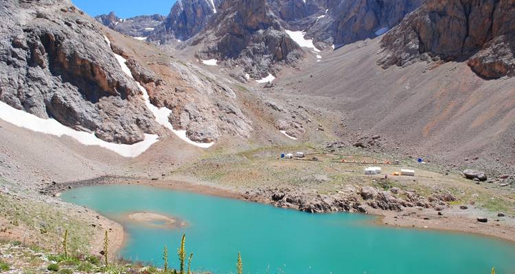 A vivid turquoise alpine lake sits amid rocky mountain walls with small expedition tents on its shore.