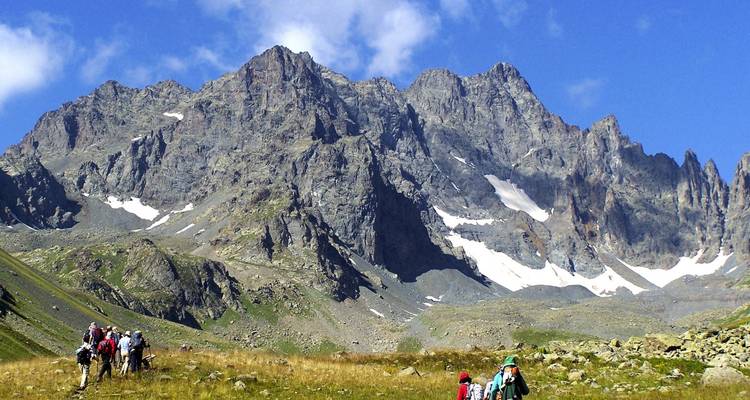 Trekkers walk through a grassy alpine basin beneath towering craggy mountains with lingering snowfields.