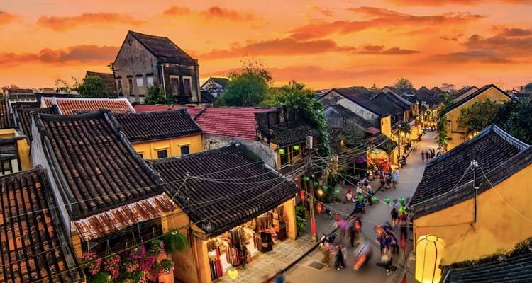 Vue surélevée d'une rue éclairée par des lanternes dans une ville historique avec des toits en tuiles sous un ciel au coucher de soleil orange vif, avec des piétons flous en bas.