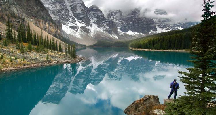 Randonneur solitaire contemplant un lac glaciaire turquoise avec des reflets de montagnes spectaculaires sous des nuages chargés.