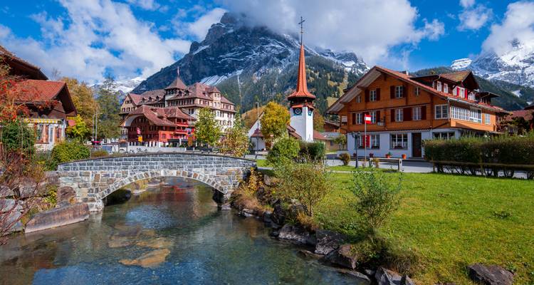 Picturesque Swiss village with stone bridge, stream and snow-capped mountains behind