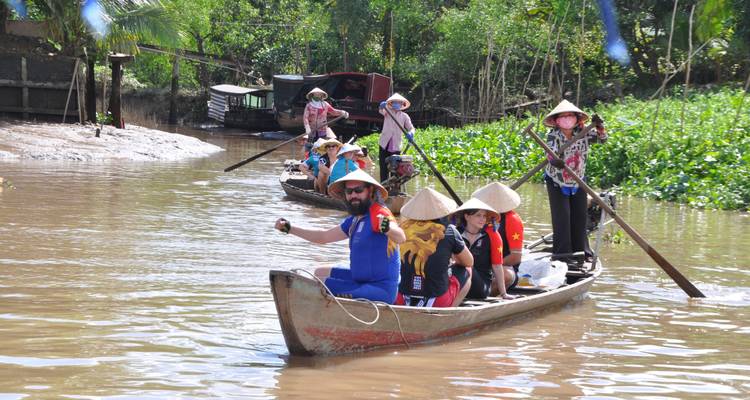 Menschen in traditionellen Hüten rudern ein Holzboot durch eine üppig grüne Wasserstraße.