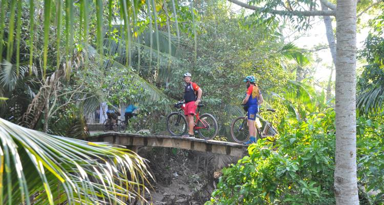 Radfahrer stehen auf einer kleinen Brücke über einem Bach, umgeben von Grün.