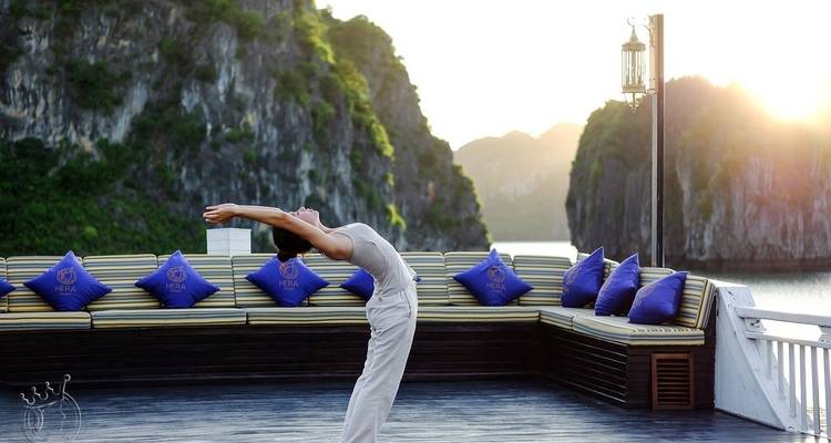Eine Person, die Yoga auf einem Bootsdeck mit dramatischen Felskulissen im Hintergrund praktiziert.