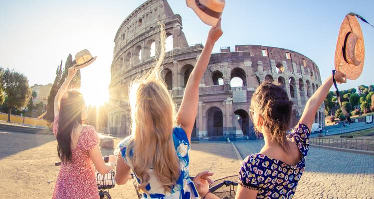 Trois jeunes femmes lèvent leurs chapeaux en signe de célébration devant le Colisée éclairé par le chaud soleil matinal.