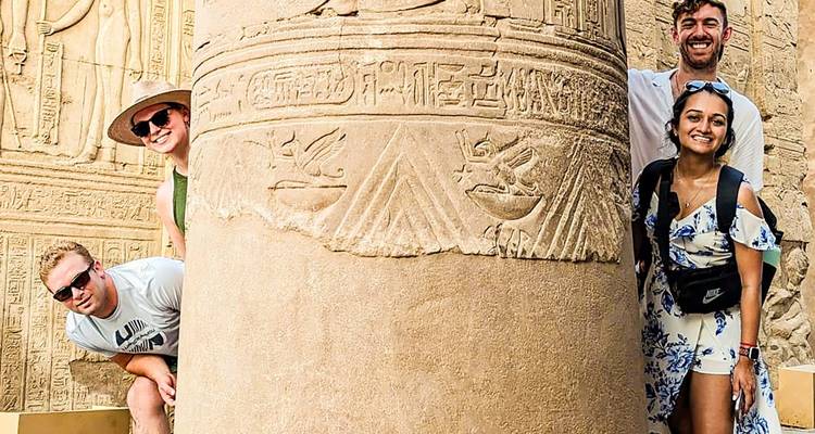 Four travellers peek playfully from behind a hieroglyph-covered column at an ancient Egyptian temple.