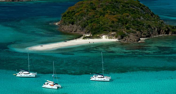Small island surrounded by crystal clear blue water with sailboats.