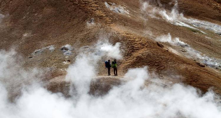 Twee trekkersers die tussen stoomende geothermische bronnen staan met witte dampclouds om hen heen.