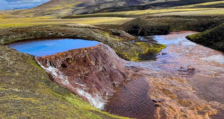 Levendig blauw warm bronwater dat over de rand stroomt naar een roestkleurig beekje in een moerig bergdal.