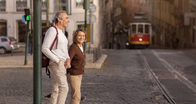 Pareja mayor sonriendo caminando de la mano en una calle empedrada de Lisboa con un tranvía amarillo clásico de fondo.